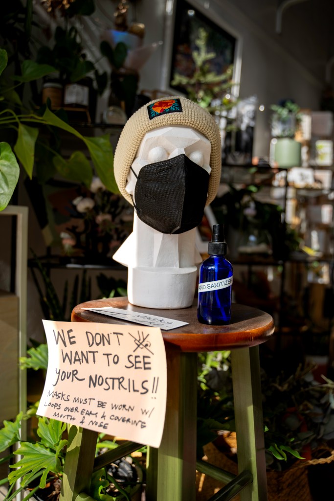 A mannequin head sporting one of the handknit hats with a golden poppy on it that Bay-Made sells. The mannequin is also wearing a KN95 mask. Next to it is hand sanitizer and a sign saying “Masks Here” which points to a basket of masks out of the frame. The sign below it says “We don’t want to see your nostrils! Masks must be worn w/ loops over ears & covering mouth and nose.” There is a little drawing of a nose with an X through it. Photo by Brian Ringo, 2023.