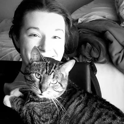 A black and white photo of a light-skinned woman leaning back in her wheelchair, from her shoulders up. Her cat is laying across her chest, looking at the camera. She is smiling.