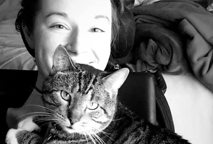 A black and white photo of a light-skinned woman leaning back in her wheelchair, from her shoulders up. Her cat is laying across her chest, looking at the camera. She is smiling.