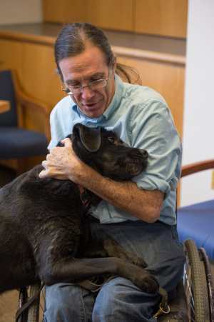 Bill Peace, a white man wearing glasses and long hair pulled back in a pony tail. He is wearing a light denim shirt looking down with affection at a black labrador dog nuzzling at his lap. He is in a manual wheelchair.