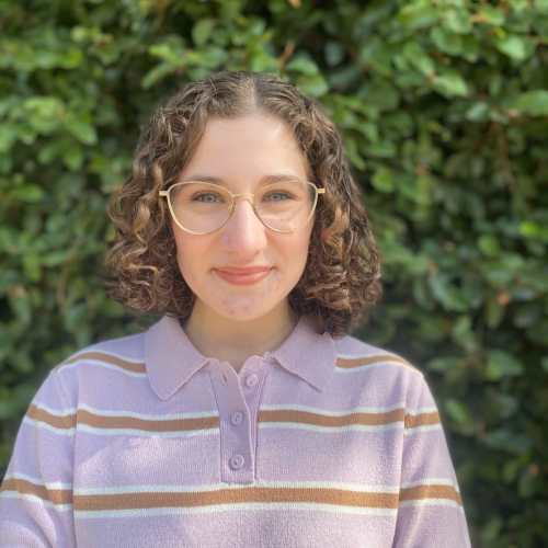 Headshot of Sabrina, a young white woman with curly, shoulder-length brown hair and glasses, wearing a purple striped sweater. Greenery is visible behind her.