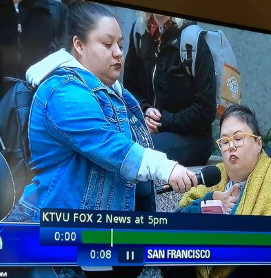Photo showing a news segment from December 16, 2019 on KTVU Fox 2 featuring Dolores Tejada holding a microphone in front of Stacey Milbern as she reads a speech during a protest in front of PG&E headquarters