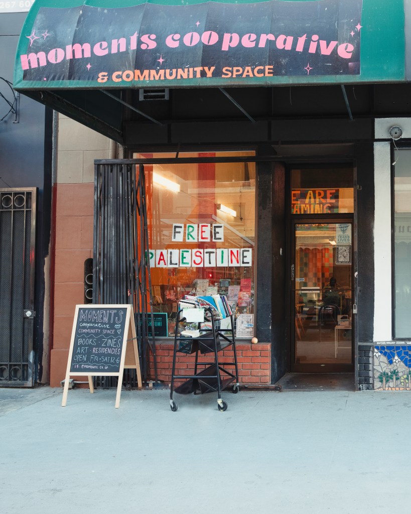 Awning reading “moments cooperative & community space” shades the store front. Beneath it, there is a cart of books for sale next to a chalkboard A-frame that reads “Moments Cooperative and Community Space. Books. Zines. Art. Residencies. Open Fri + Sat 12-6. Ig: @moments.co.op.” In the window are more books and the words “FREE PALESTINE.” The door includes small prints reading “Black Trans Lives Matter.” A warm glow of light emanates from the cozy bookshop within. Photo by Brian Ringo, 2023.