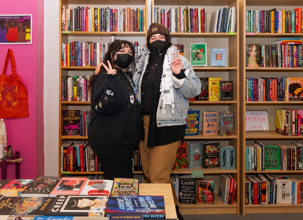 Samantha and D, Moments Cooperative members, pose in front of the store’s shelves full of books for sale. They are both wearing black KN95 masks and making peace signs while posing. In front of them is another table full of featured books, including Creep by Myriam Gurba, Horse Barbie by Geena Rocero, and Africa Risen, a speculative fiction anthology by African and African diaspora writers Edited by Sheree Renée Thomas, Oghenechovwe Donald Ekpeki, and Zelda Knight, among other books. Photo by Brian Ringo, 2023.