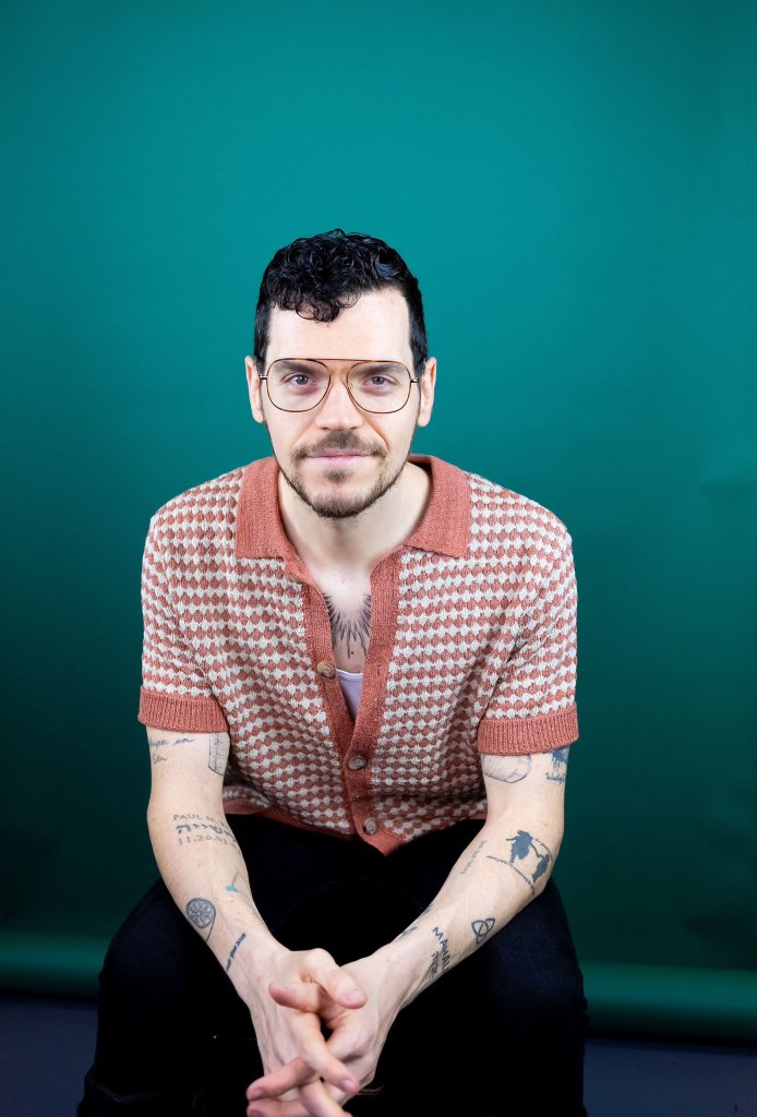 A white man with curly dark hair, glasses, and tattoos on his arms and chest, wearing an orange and white patterned shirt with black pants. He’s sitting down in front of a dark green background.