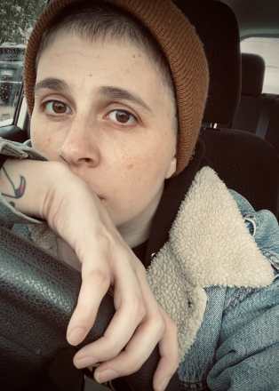 A Andrews, a white non-binary person with brown eyes and short hair. They are sitting in the driver’s seat of a car with dark seats, wearing a tan beanie, and lined denim jacket.