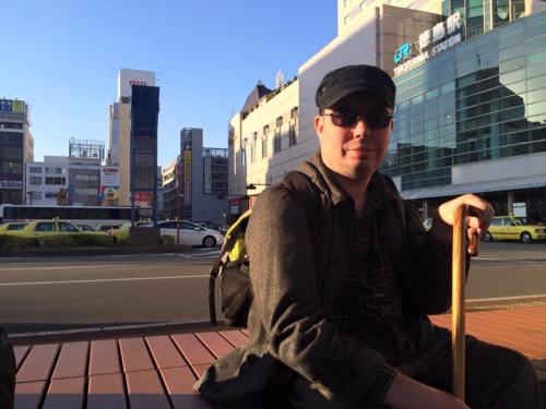 A white man sitting on a bench with a cane. He is wearing dark sunglasses and has a black cap on his head. Behind him is an urban landscape of a city in Japan