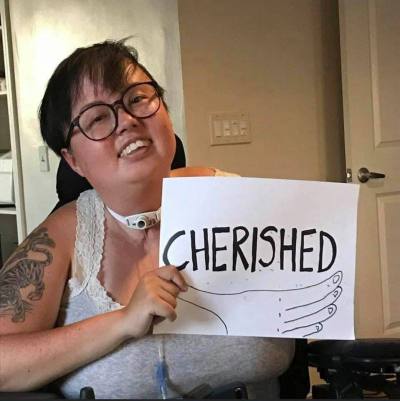 A photo of Stacey Park Milbern, a mixed race Korean and white queer person, holding a paper sign that reads “Cherished.” The photo is stylistically torn at the edges with cursive text appearing across the bottom, “Stacey Milbern.”