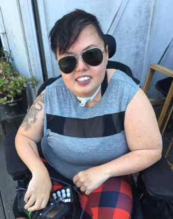 Stacey Milbern Park outside smiling at the camera with gold rimmed aviator sunglasses on. Her short, undercut styled hair is swooped forward over her forehead a little to the right. A tiger tattoo curls up her bare right arm. She is wearing a grey sleeveless tee shirt with a bold black horizontal stripe and red and black plaid leggings.