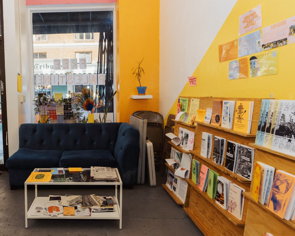 The cozy zine corner of Moments is painted yellow with zines lining the wall next to a small blue couch. Extra tables and chairs are stacked in the corner. A small coffee table is in front of the couch covered in zines. Behind the couch is the window loo looking out onto 13th street, where part of the Oakland Tribune sign is visible across the street. Photo by Brian Ringo, 2023.