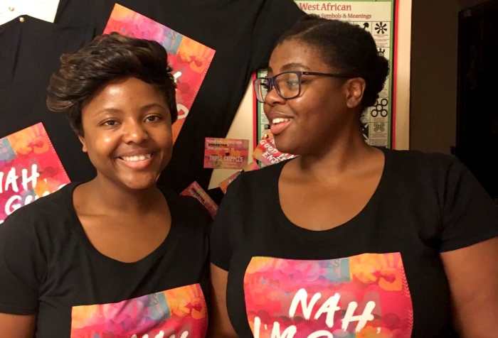 A picture of two Femmes/Women with almond brown skin and black hair, one with glasses and a ponytail, the other with a short cropped hairstyle. Each of them is wearing a black T-shirt with a colourful slogan saying “Nah, I’m good love. Enjoy.” In the background, the same T-shirts hang behind them on a cream wall, next to a poster with West African adinkra symbols.