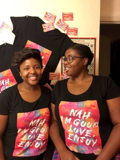 A picture of two Femmes/Women with almond brown skin and black hair, one with glasses and a ponytail, the other with a short cropped hairstyle. Each of them is wearing a black T-shirt with a colourful slogan saying “Nah, I’m good love. Enjoy.” In the background, the same T-shirts hang behind them on a cream wall, next to a poster with West African adinkra symbols.