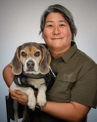 Photograph of Karen Nakamura (an asian american woman with short hair) holding her service dog Momo, a grey and white beagle. The photograph is studio quality.