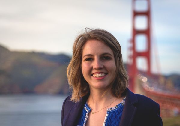 Image description: a young white woman with shoulder-length blonde hair standing outdoors with the Golden Gate Bridge and the Marin Headlands behind her. She is wearing a navy blazer with a blue sweater inside.