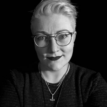 A black-and-white headshot of white person with short platinum blond hair and light eyes. They are wearing a dark grey sweater, round wire-framed eyeglasses, and dark lipstick, and are smiling slightly at the camera.