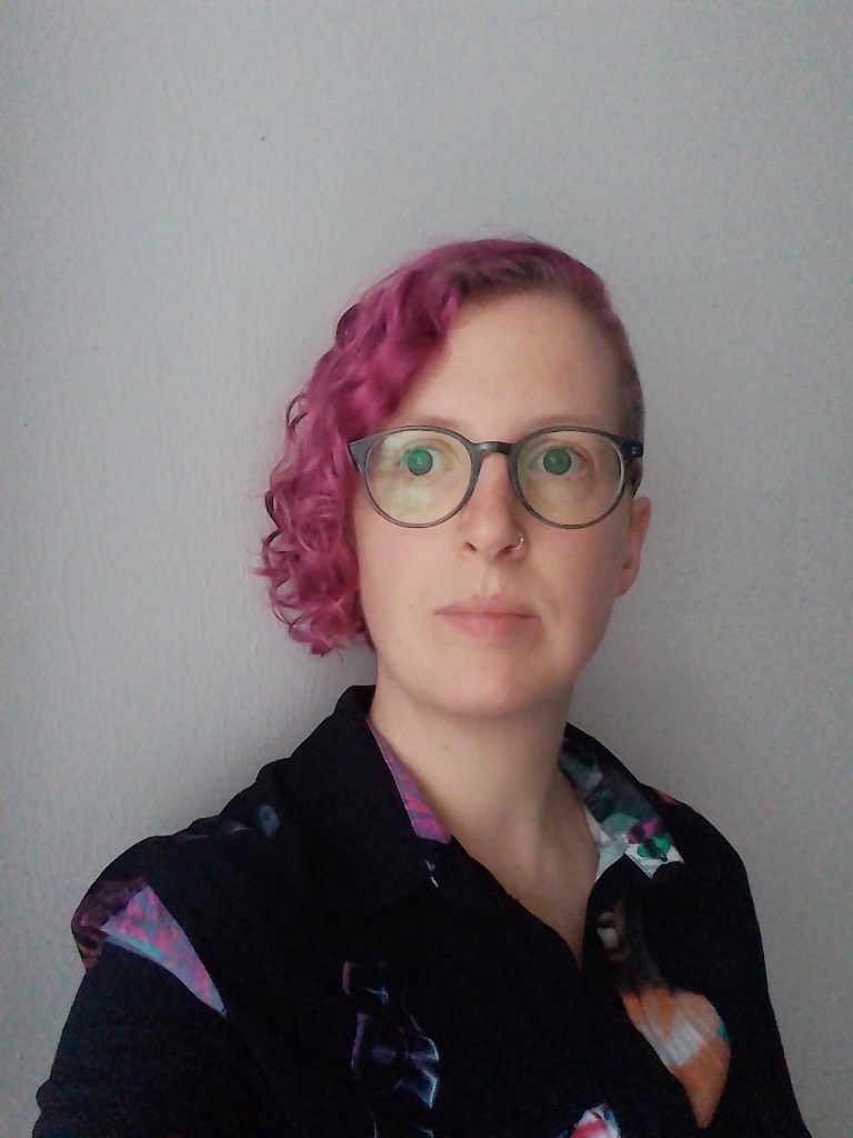 A photo of a white woman against a grey wall, her head is tilted slightly to the right, looking away from the camera. She has curly hair, dyed magenta, uses greenish glasses and is wearing a black shirt.
