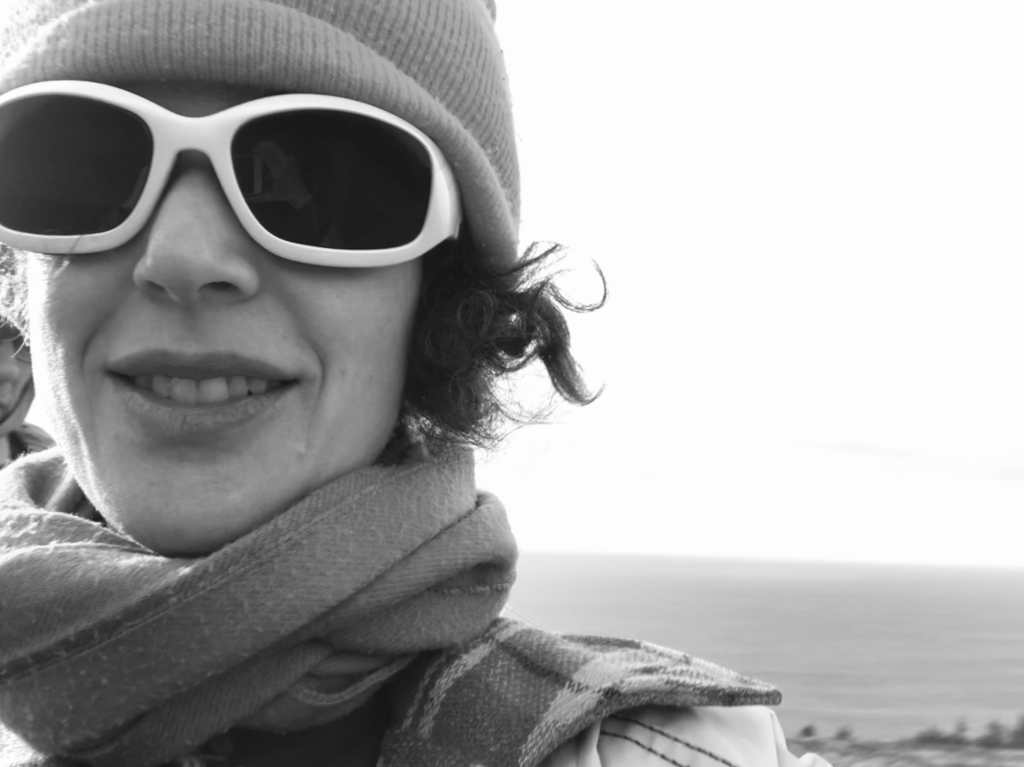 A selfie of Aislinn in front of the ocean on a winter day. She is a white person wearing a toque, scarf, and big, dark, somewhat bug-like sunglasses.
