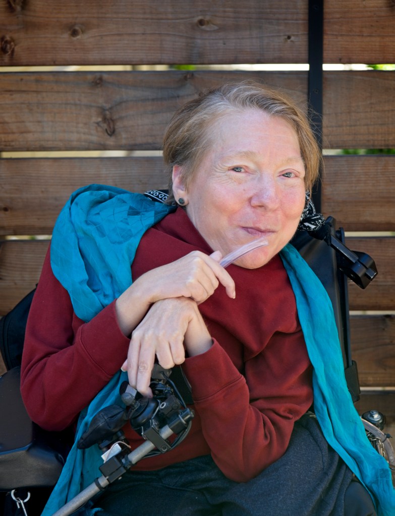 Alana Theriault, a white woman, is sitting in a power wheelchair and smiling. She has short reddish-blonde hair that is beginning to gray. She is wearing a rust colored long sleeve shirt, a blue scarf, and gray slacks, and is holding a narrow clear ventilator tube in her hand. The background is a wooden fence. Photo Credit: DeAnna Tibbs Photography (deannatibbs.com) 