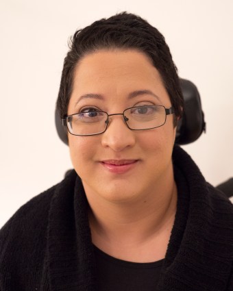 A headshot-style photo of Alejandra Ospina, a Latin American woman with short, dark hair and brown eyes. She is wearing a black top and an open black sweater. She is smiling slightly with her mouth closed, and she is wearing black-framed glasses. 