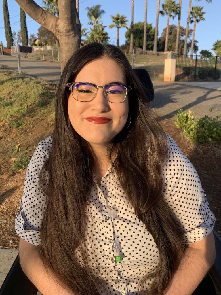 Alena Morales, a Mexican Filipina woman, sitting in her wheelchair among San Diego palm trees. Her long brown hair is styled down around her shoulders, and she dons red lipstick with winged eyeliner. She is wearing a white button up shirt with black polka dots, and her central line hangs out from the neckline of her shirt.