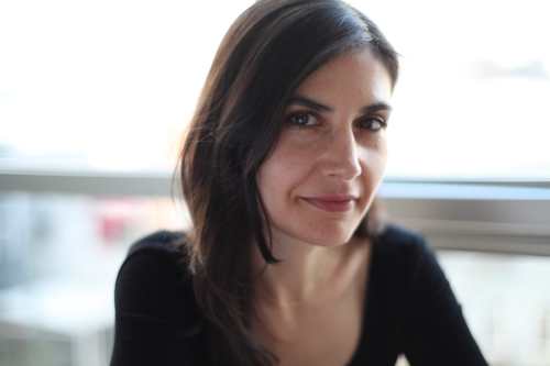 Alexis, a white woman with long dark hair, sits inside in front of a window. She is wearing a long sleeved black shirt and smiles softly at the camera.