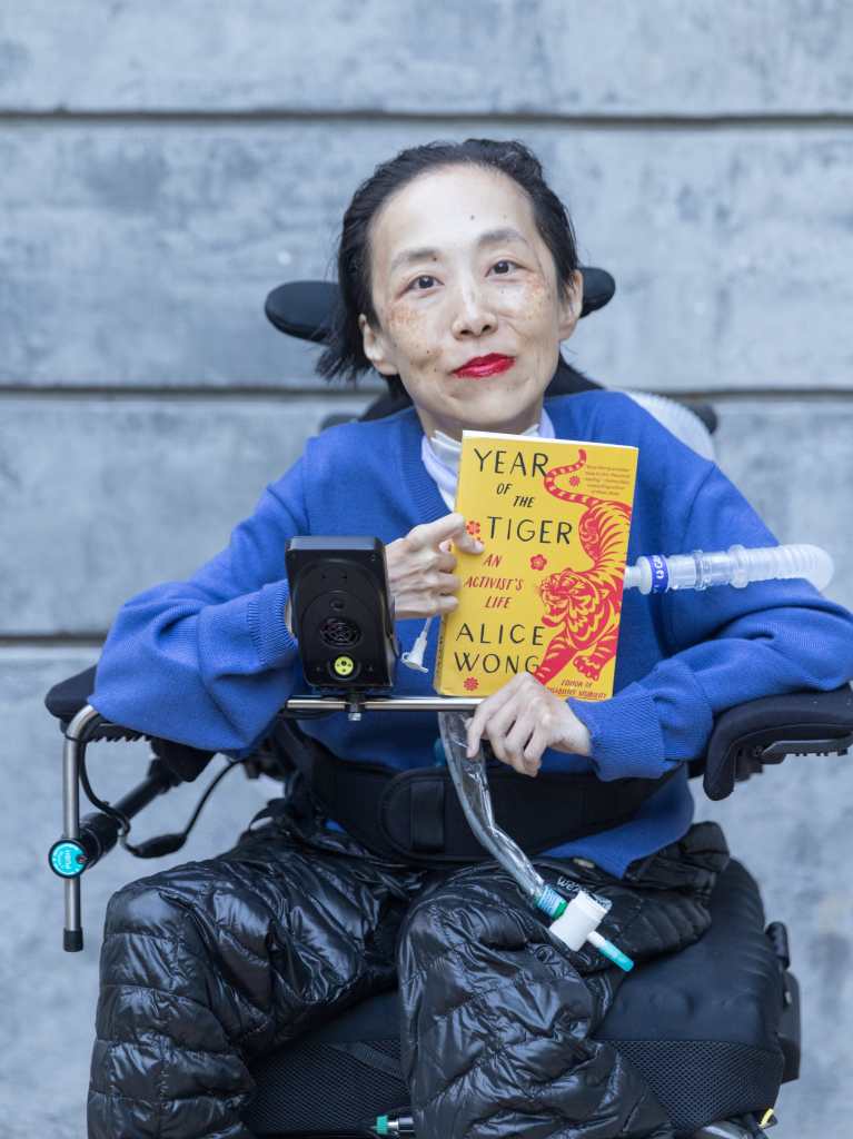 Photo of Alice Wong, an Asian American disabled woman in a power chair. Behind her is a gray concrete wall. She is wearing a bold red lip color, blue cardigan and black puffy pants. A trach and ventilator tube is at her neck and shoulder. She is holding a copy of her memoir, YEAR OF THE TIGER, a yellow book cover with a fierce tiger and red flowers with the text after the title, An Activist’s Life, Alice Wong, Editor of Disability Visibility. Photo credit: Eddie Hernandez Photography.
