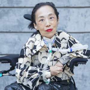 Photo of Alice Wong, an Asian American disabled woman in a power chair. She is wearing a black blouse with a floral print, a bold red lip color and a trach at her neck. In the background is a gray cement wall. Photo credit: Eddie Hernandez Photography.