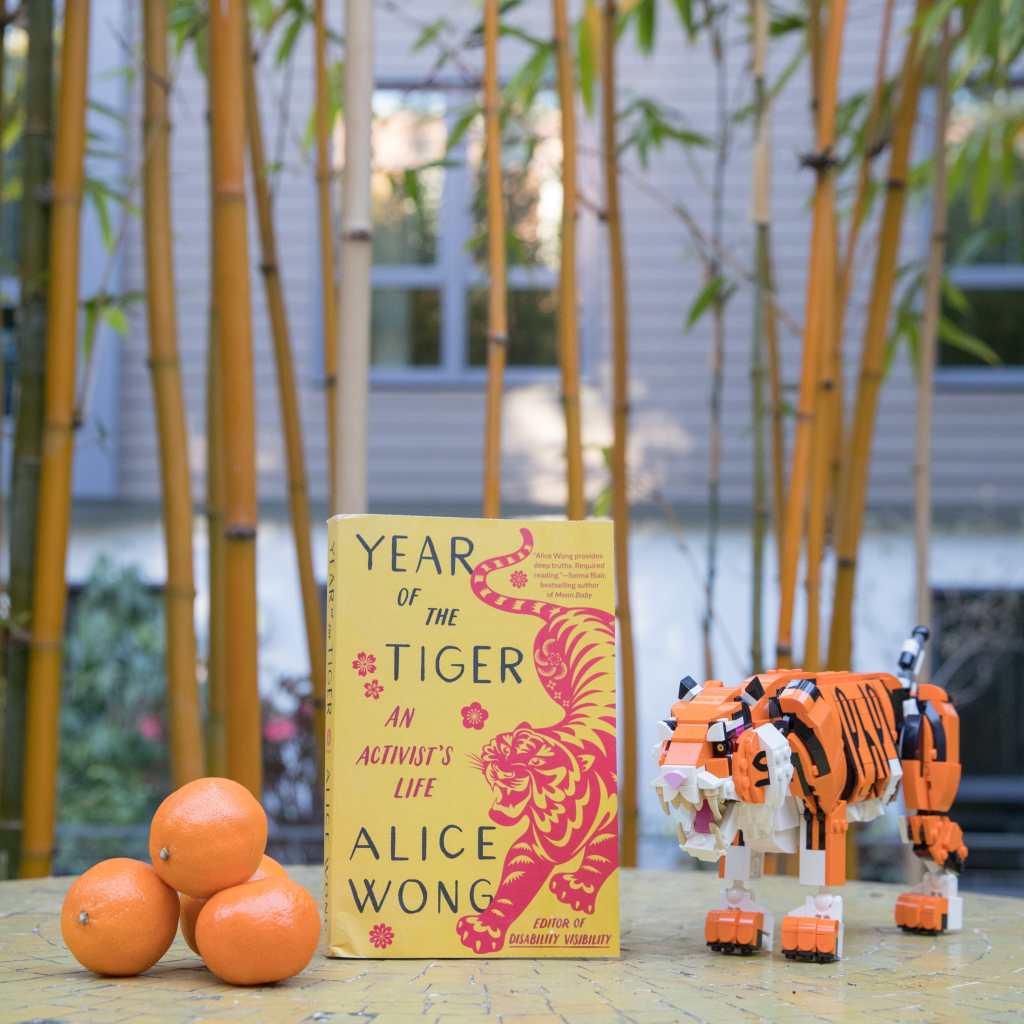 Photo of YEAR OF THE TIGER, a paperback with a marigold yellow background. On the right side is an illustration of a crouching tiger in red in the style of Chinese paper cuttings with delicate cutouts in various shapes giving form and definition to the tiger. The tiger has a fierce expression, eyes and jaws wide open, teeth bared. The tiger has large paws with four claws extended. On the left in black large text YEAR OF THE TIGER at the top and ALICE WONG below. In the center in smaller red text AN ACTIVIST’S LIFE and in the lower right corner EDITOR OF DISABILITY VISIBILITY. Small, delicate red flowers are sprinkled throughout. Book cover by Madeline Partner. Next to the book is a small pyramid of tangerines and to the left is a Lego figure of a fierce tiger. In the background are bamboo trees. Photo credit: Eddie Hernandez Photography.