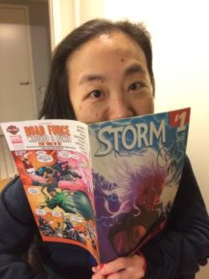 Asian American woman in a wheelchair wearing a navy hoodie. She is holding a comic book by Marvel, Storm #1, close to her face, covering her mouth. 