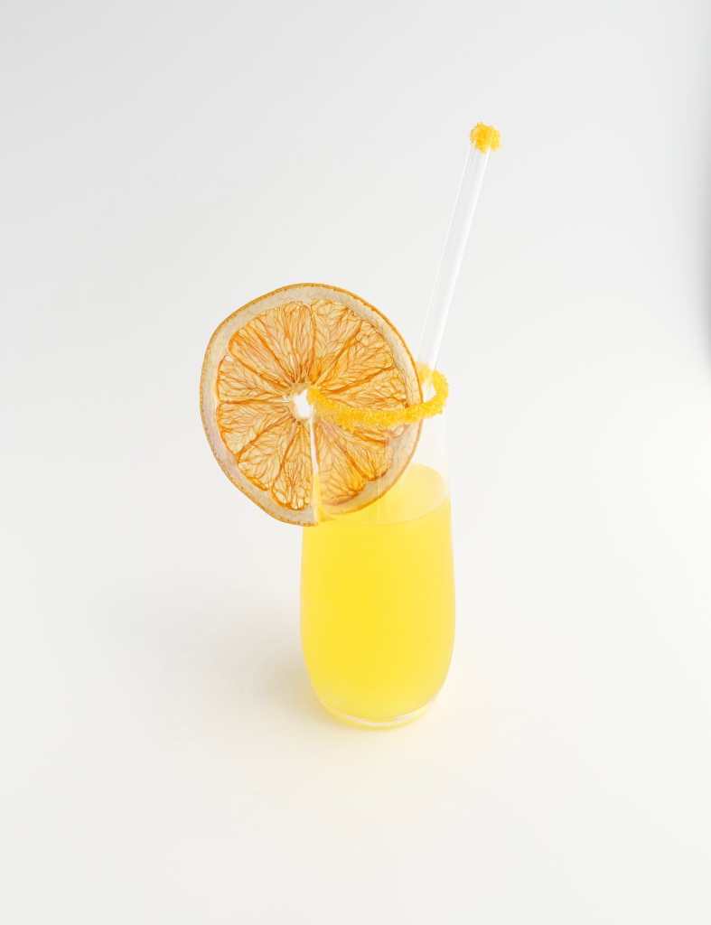 A stemless Champagne flute filled with a bright yellow cocktail, with a golden yellow sugar rim, a glass straw with golden yellow sugar on top, and a large, dehydrated grapefruit wheel sitting on the rim of the glass.
