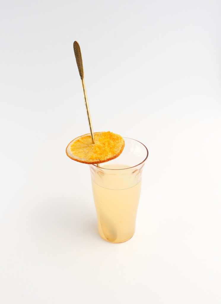 A small, thin pink cocktail glass filled with a pale yellow cocktail, with a gold stirring spoon and a dehydrated orange wheel through it, with half of it covered in golden yellow sugar.