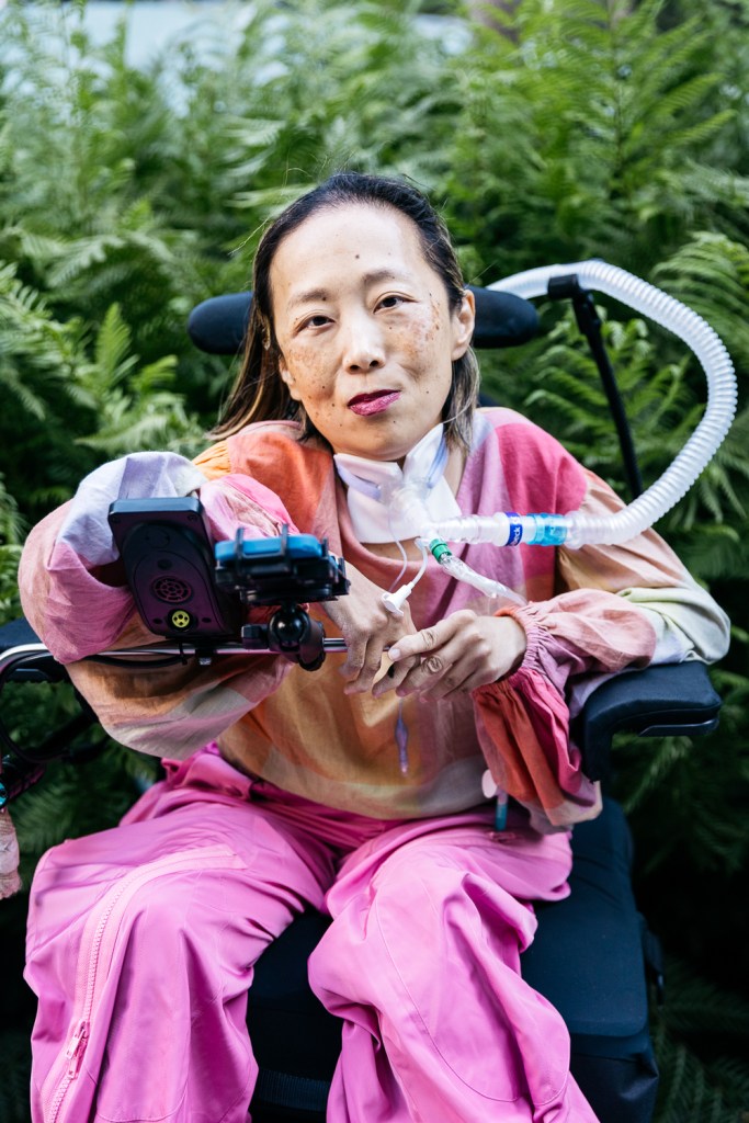 Alice Wong, Asian American woman in a wheelchair with a tracheostomy at her neck connected to a ventilator. She’s wearing a pink plaid shirt, pink pants, and a magenta lip color. She is smiling and behind her are a bunch of large fern-like plants. Photo credit: Allison Busch Photography.