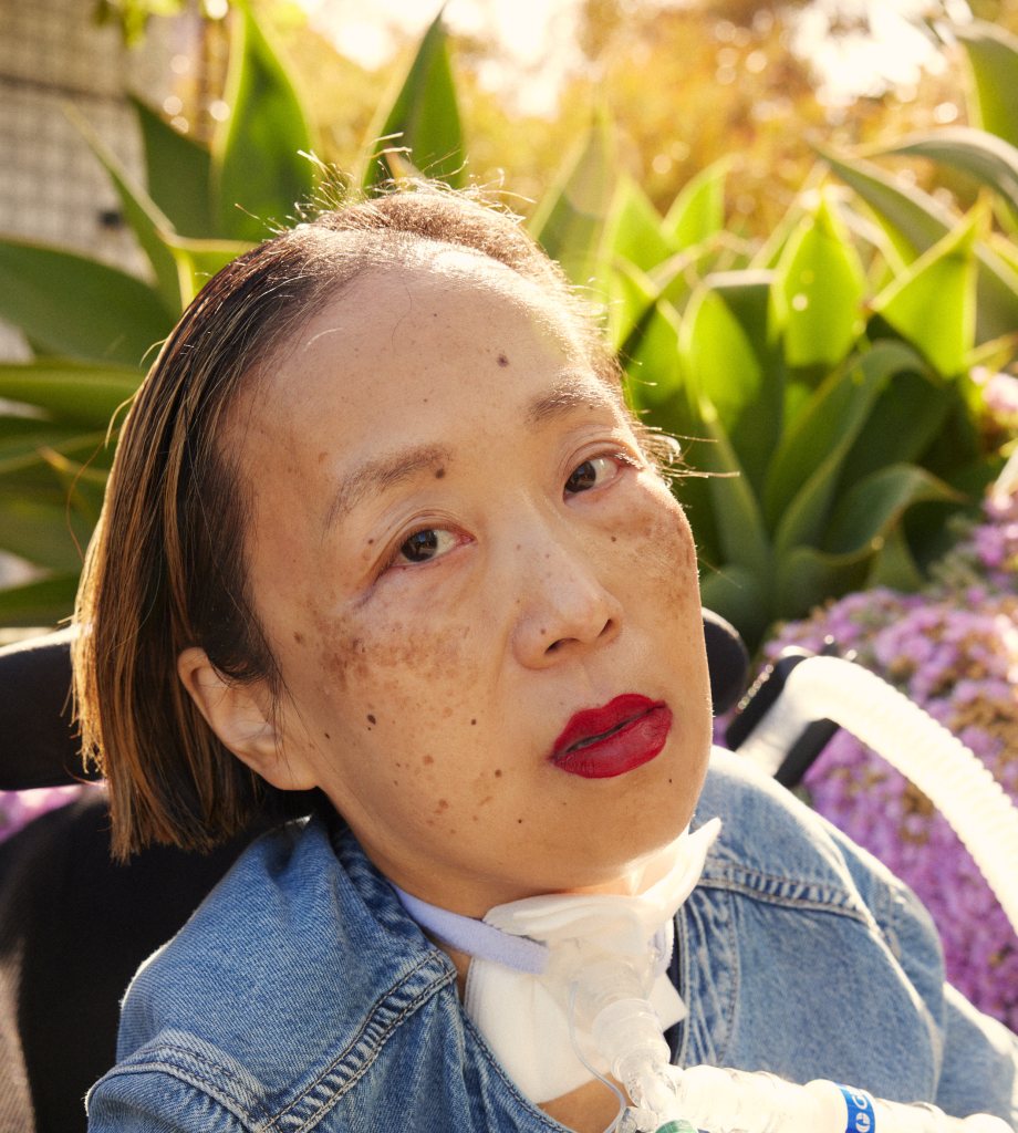 lice Wong, an Asian American disabled woman with a tracheostomy at her neck. She is wearing a bright red lip color and a denim shirt. She is looking intently into the camera and the sun is shining behind her with a bunch of plants. Photo credit: María del Río