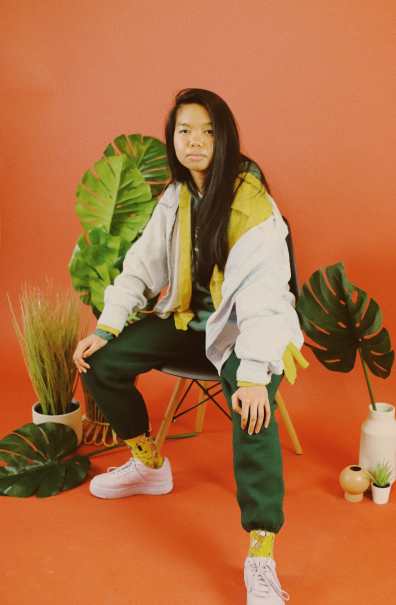 Allison Masangkay (aka DJ Phenohype), a Filipinx femme, sits in a chair wearing an outfit of yellows, greens, lavender, and light wash denim blue with a warm orange backdrop, surrounded by several green plants. They look directly at the camera with a serious expression and their hands resting on their knees. Photo by Bianca Recuenco.