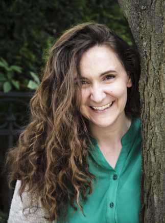 A brown-haired brown-eyed woman stands smiling beside a tree trunk. She wears a deep green shirt and an ivory cardigan.