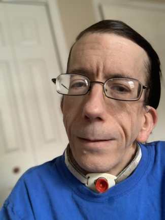 Andrew Pulrang, a white, clean-shaven man with glasses and black hair, wearing a blue shirt. A tracheostomy is visible around his neck just above the shirt collar.