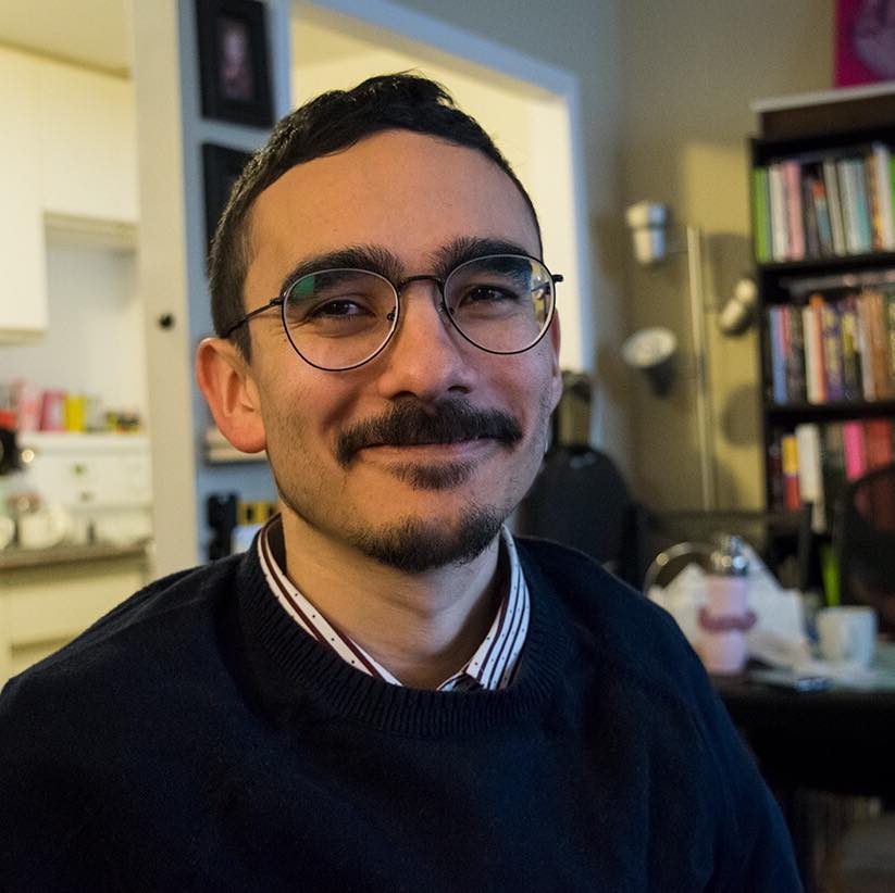 A close-up photo of Angelo, a man smiling with dark metal glasses and a short beard. He is wearing a navy blue sweater layered over a striped collared shirt.