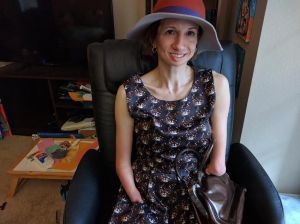 This is a photograph of an amputee woman, wearing an orange and tan hat and a blue dress with a print of brown, grey and white cat faces on it. She is seated in a chair and holds a brown leather purse on her lap. Her right art ends with a pointed tip, and her left arm, shorter than the right one, ends with a more rounded “hand” with a fleshy ball on it. She looks at the camera with a large smile.