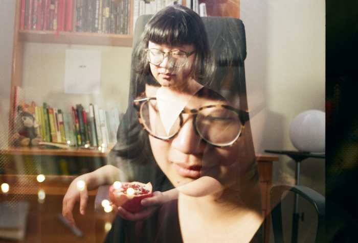 A double exposure photograph, wherein two images are faded onto one another in a single image, of Jane sitting at her desk on a black office chair.  Jane is a light skin East Asian person, with her eyes closed, wearing brown and black glasses and short black hair that slightly flares at the end with rounded bangs like a bowl. In one exposure Jane’s face is larger and closer to the camera; there are lights directly behind her. In the other, she is sitting on a black office chair and holding a sliced open half of a pomegranate. Behind her is a round, moon-like light. The desk behind her is a medium orange-brown and contains a head shelf, with books ordered in red, black, white, and the rest of the rainbow ordered on the top of the table. There’s a small brown monkey plushie near the left end of the desk. There’s a black and white ink illustration above the orange to blue books on top of the desk where there’s an open space.