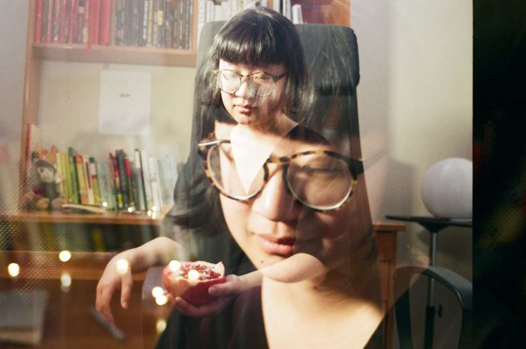 A double exposure photograph, wherein two images are faded onto one another in a single image, of Jane sitting at her desk on a black office chair.  Jane is a light skin East Asian person, with her eyes closed, wearing brown and black glasses and short black hair that slightly flares at the end with rounded bangs like a bowl. In one exposure Jane’s face is larger and closer to the camera; there are lights directly behind her. In the other, she is sitting on a black office chair and holding a sliced open half of a pomegranate. Behind her is a round, moon-like light. The desk behind her is a medium orange-brown and contains a head shelf, with books ordered in red, black, white, and the rest of the rainbow ordered on the top of the table. There’s a small brown monkey plushie near the left end of the desk. There’s a black and white ink illustration above the orange to blue books on top of the desk where there’s an open space.