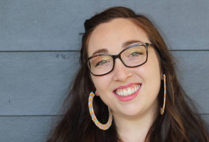 A pale white woman with brown hair, green eyes, and tortoiseshell glasses leans against the blue siding of a house, looking at the camera and smiling. She is in a blue, denim shirt and has on large blue and yellow beaded hoop earrings and a black and gold Magen David necklace.
