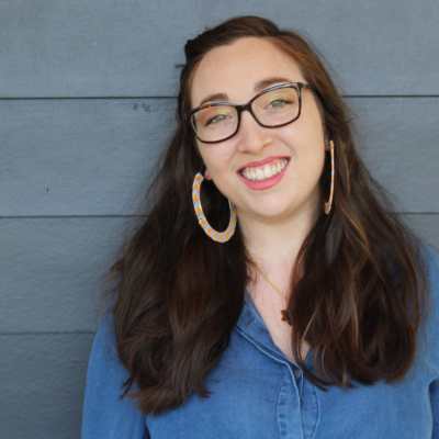 A pale white woman with brown hair, green eyes, and tortoiseshell glasses leans against the blue siding of a house, looking at the camera and smiling. She is in a blue, denim shirt and has on large blue and yellow beaded hoop earrings and a black and gold Magen David necklace.