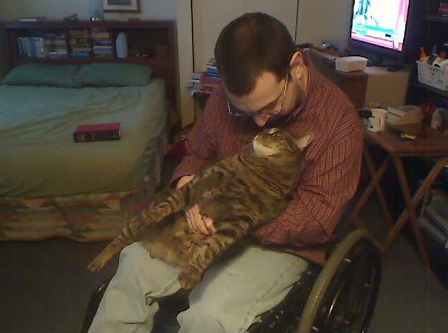 Image of a white man in a wheelchair. His head is bent down toward the face of his cat, a Maine coon, who is sitting on his lap.