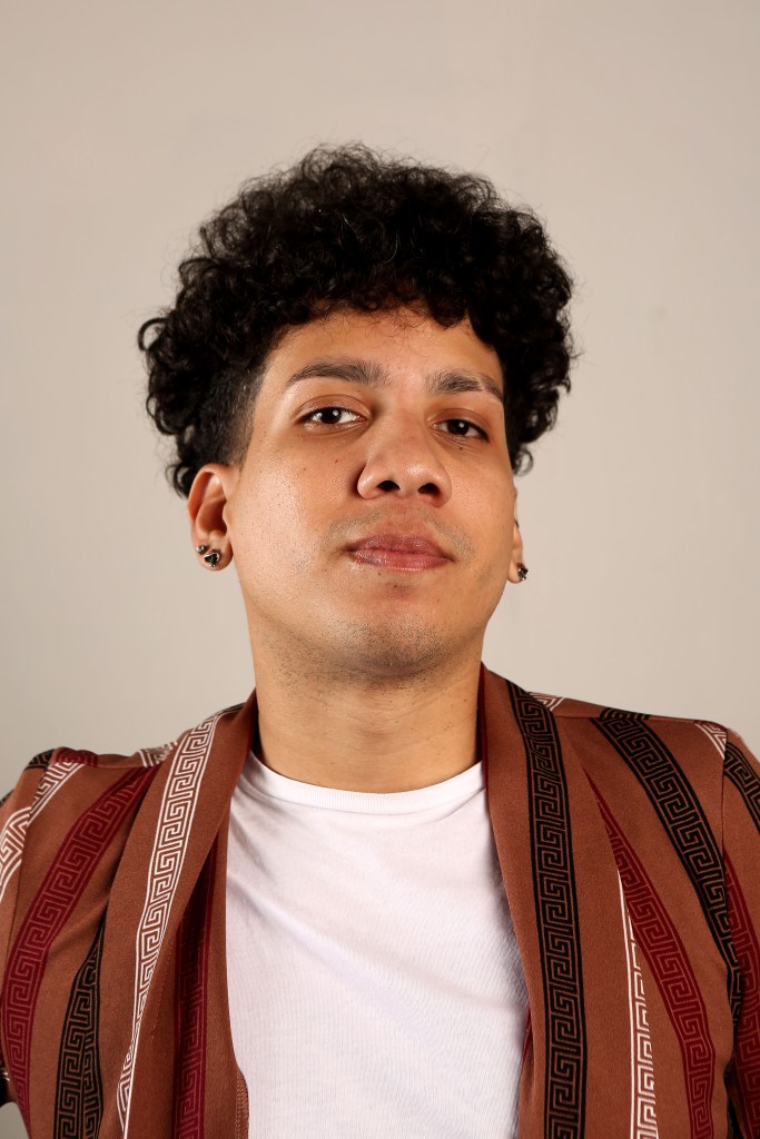A photo of Ínaru, a non-binary black person that has brown colored skin, big lips, big cheeks, upturned eyes, wide nose and ears holding fungus stud earrings. Their haircut is a small afro and they are wearing a white shirt with a brown-colored cardigan that has black, velvet and white lines that run from the upper to the latter part of the cloth