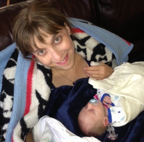 A young white woman with brown hair wrapped in a blanket with her newborn baby in front of her.