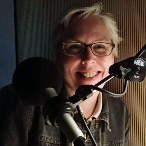 A white woman with short white hair inside a recording booth. There is a microphone in front of her and light from the corner illuminating the right-half of her face. She is wearing a dark-colored long-sleeve shirt and eyeglasses. She is smiling at the camera. 
