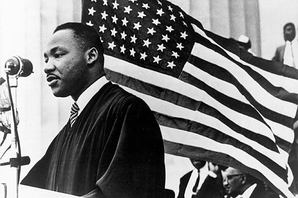 A black-and-white image of an African-American man speaking at a lectern. He is facing a crowd and standing in front of a large American flag.
