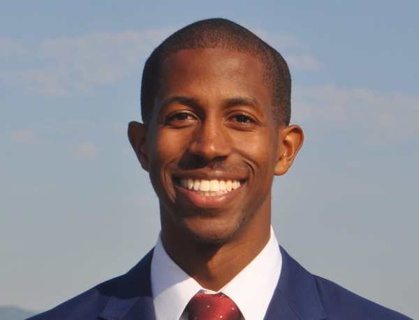 Dr. Justin Bullock, a Black man with short hair wearing a navy suit with a white shirt and dark red tie. He is smiling at the camera.