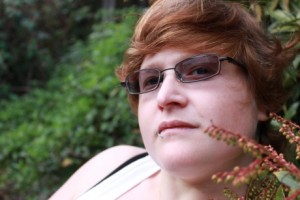 A woman with wavy red hair wearing tinted glasses staring at the camera. There are green bushes in the background.
