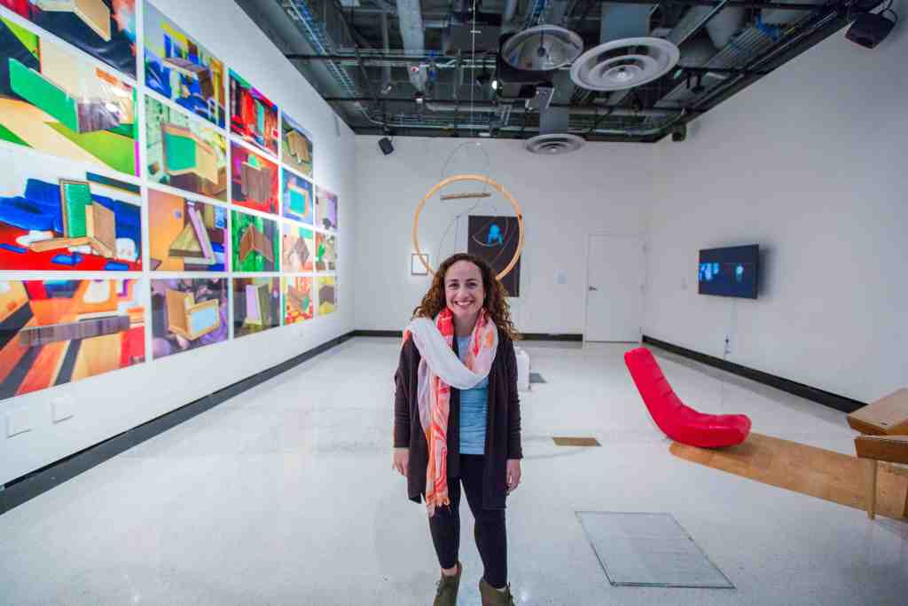 A short-statured woman with long brown curly hair and brown eyes stands in the middle of a white cube gallery space, smiling at the camera. She is wearing red lipstick to match the red and white scarf wrapped around her neck. On every wall of the gallery space hangs contemporary art that includes brightly-colored photographs of abstract shapes, a round wooden ring hanging from the ceiling, a black and white video installation of a sign language interpreter and a low-lying red lounge chair on a mat. The ceiling is exposed so all the beams, wiring and fan ducts are visible.